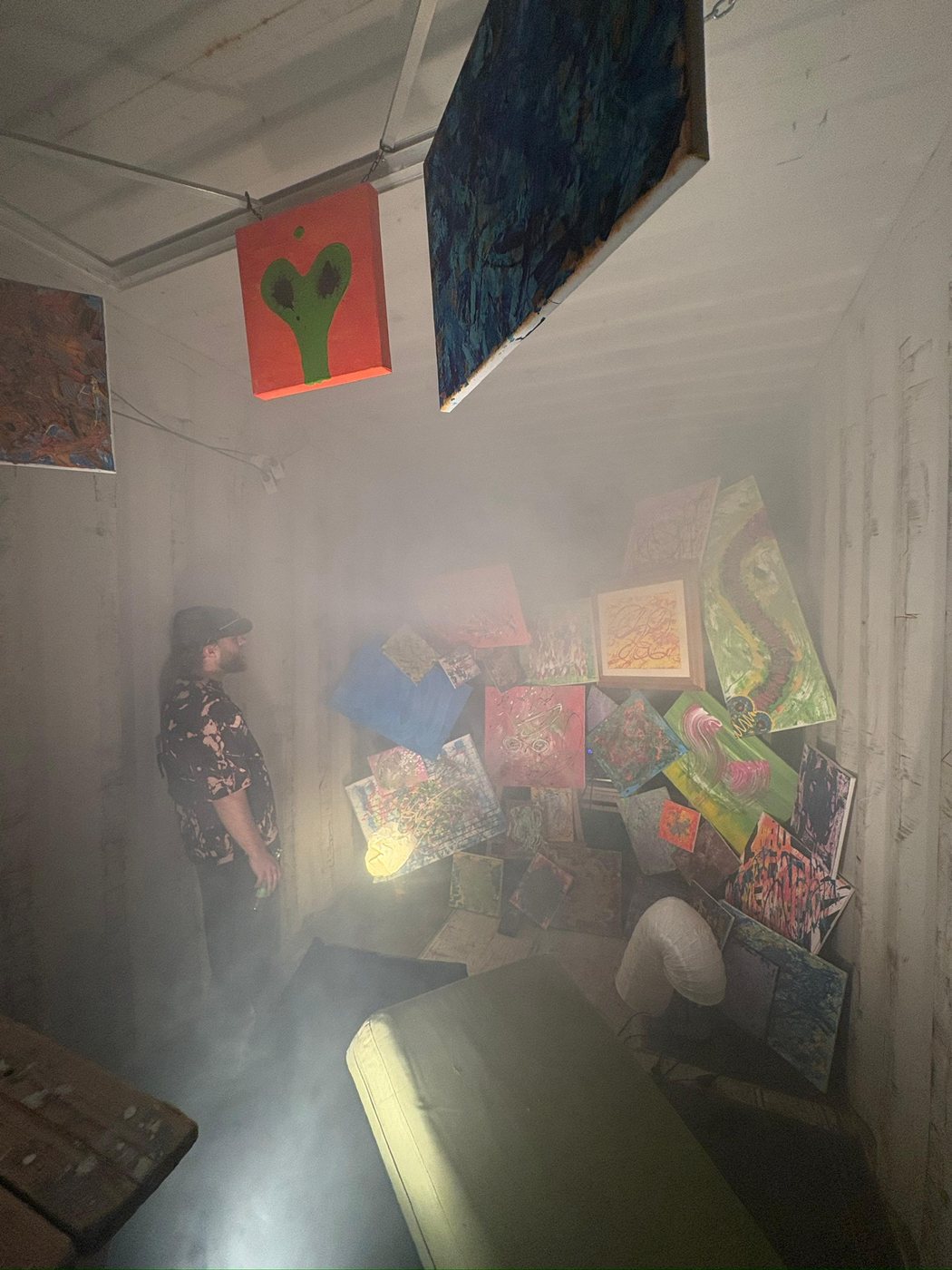 Wessel in the shipping container, the walls hung edge to edge with paintings, light cutting through the haze